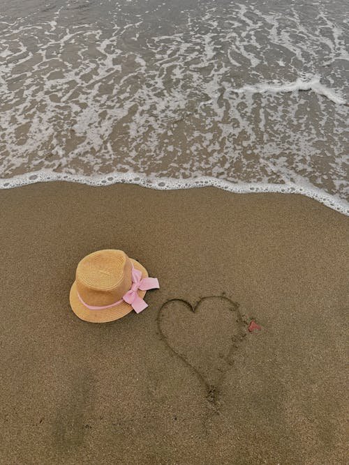 Cappello di paglia con disegno a forma di cuore posizionato sulla sabbia di una spiaggia al sole, suggerendo un accessorio per proteggersi dal sole.