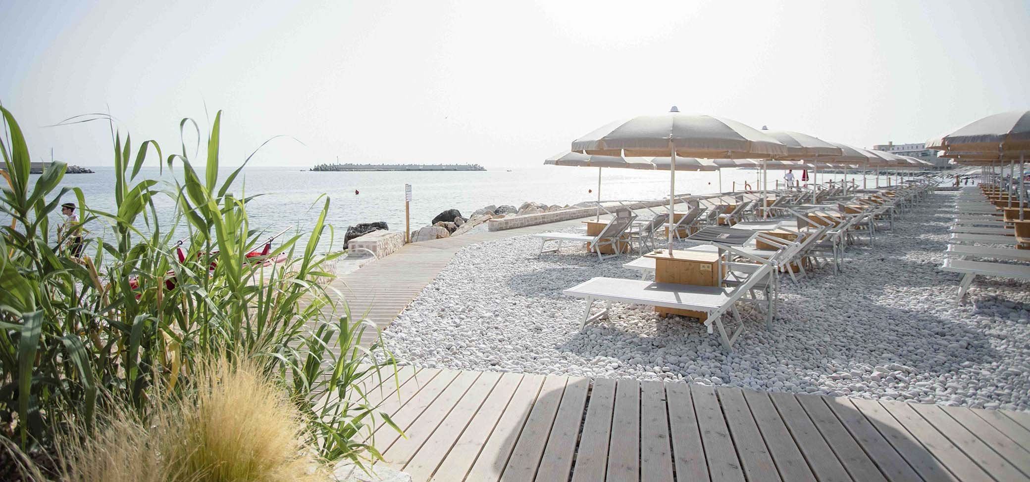 Spiaggia attrezzata con ombrelloni e lettini allineati sulla ghiaia, passerella in legno, mare calmo e cielo claro; piante verdi sul primo piano.
